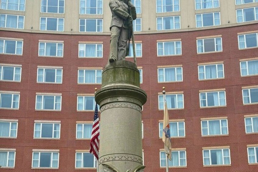 A statue on a pedestal with two flags, a red brick building with many windows in the background. 4