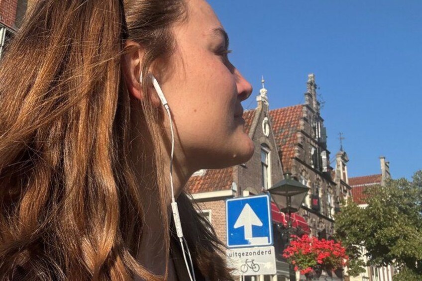 A person with long hair wearing earphones, a blue directional sign, and a building with red-tiled roofs. 4