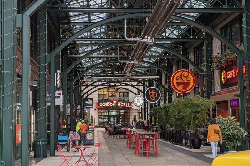 An indoor market with a glass ceiling, tables, chairs, and various shops including Gordon Hotel and Cafe Y. 4