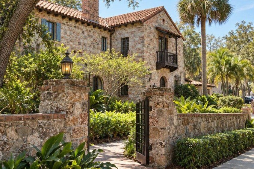 A stone house with a tiled roof, a gated entrance, and lush greenery including palm trees. 1