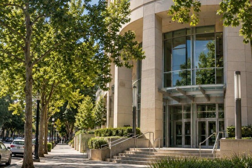 A modern building with large windows and a curved facade, surrounded by trees and a sidewalk. 1