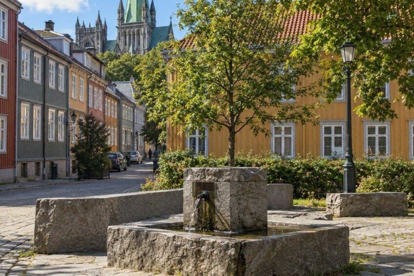 A cobblestone street with historic buildings, a tree-lined square, and a church spire in the background. 1