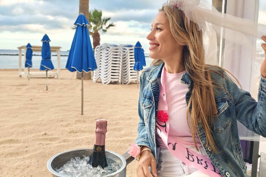 A woman sitting by a beach with a champagne bottle in a bucket. 2