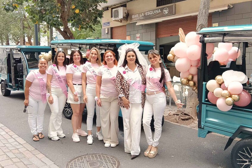 A group of people in white outfits with pink sashes, standing in front of a teal vehicle decorated with pink and gold balloons. 4