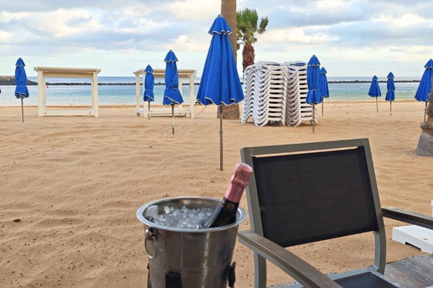 A beach with lounge chairs, blue umbrellas, and a bottle of champagne in a bucket. 3