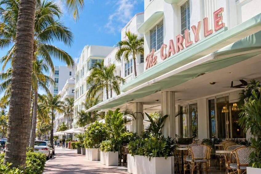 A street with palm trees, a building with 'The Carlyle' sign, outdoor seating, and a clear sky. 3