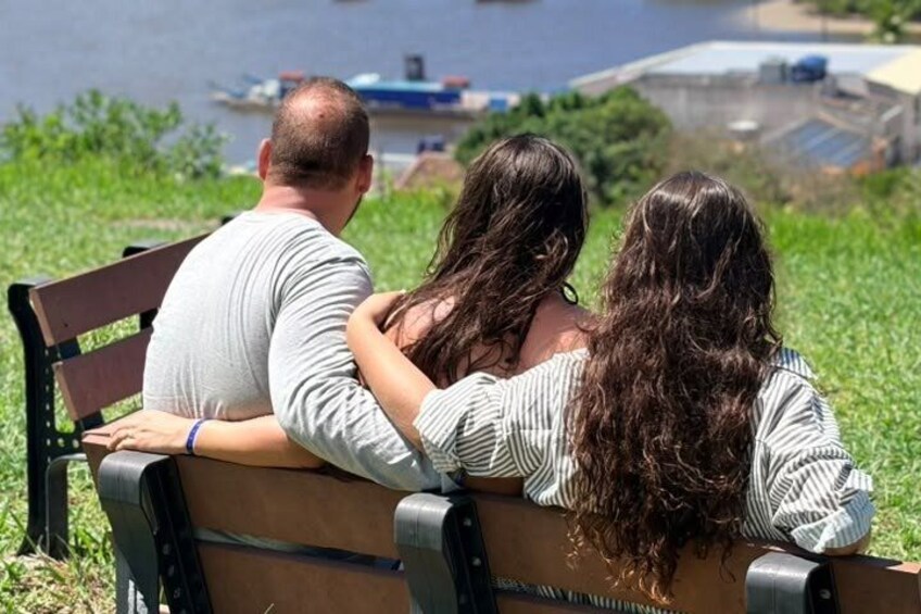 Three people sitting on a bench facing a body of water with boats and industrial structures in the background. 4