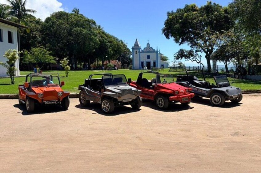 Four off-road vehicles parked in a line on a dirt surface, with a church and lush greenery in the background. 3