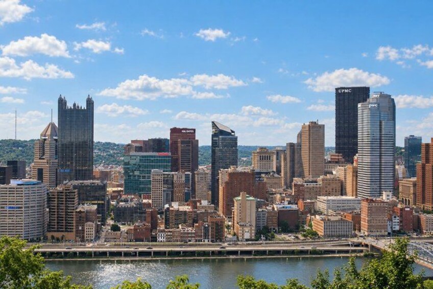 A city skyline with various buildings, including a prominent UPMC building, a river in the foreground, and a clear blue sky. 3