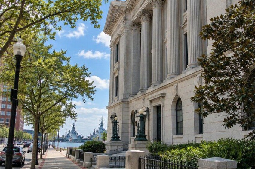 A classical building with columns, a sidewalk, trees, and a distant view of a cityscape. 5