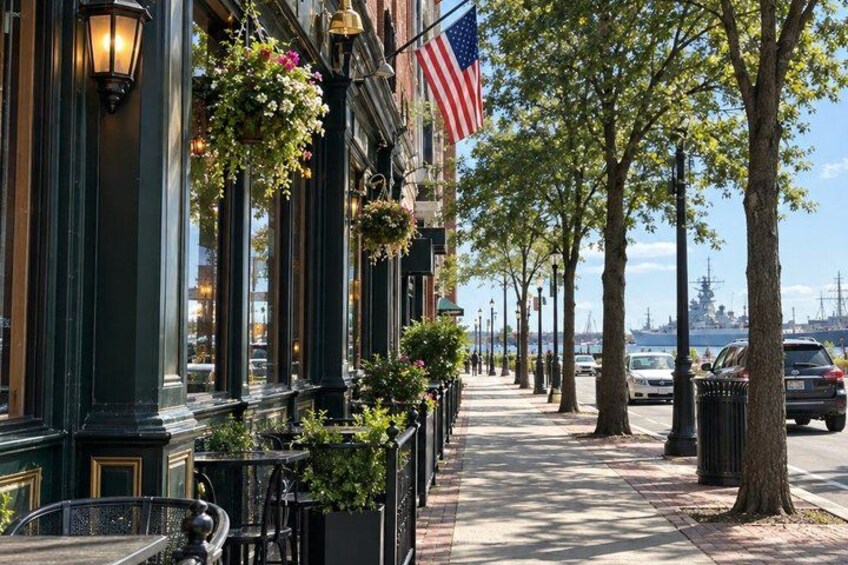 A street with a row of buildings, an American flag, hanging flower baskets, and a ship in the background. 3