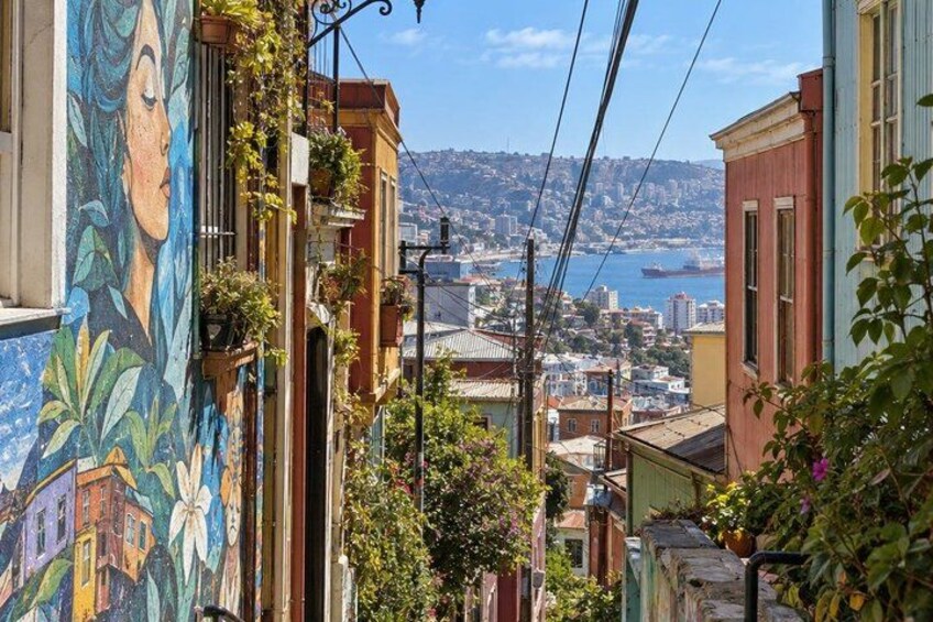 A narrow street with colorful buildings, a mural, and a view of a distant city and water. 3