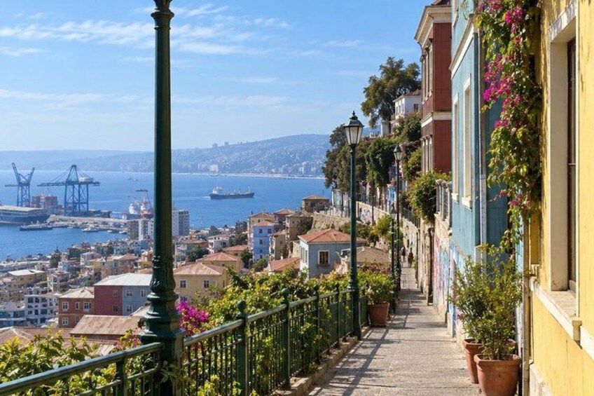 A coastal cityscape with a narrow street lined with colorful buildings, a clear blue sky, and a view of the sea and port in the distance. 5