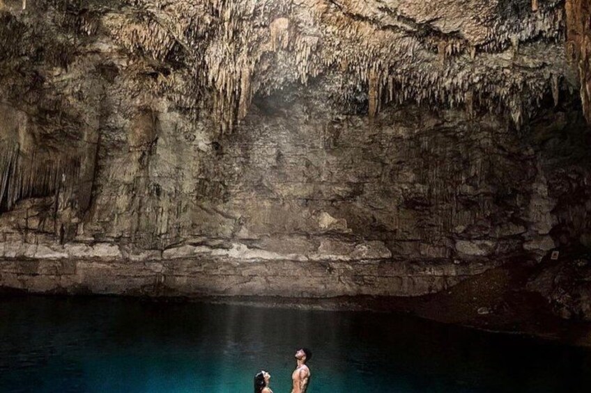 A cave with stalactites and stalagmites, a pool of water, and two people standing by the water. 2