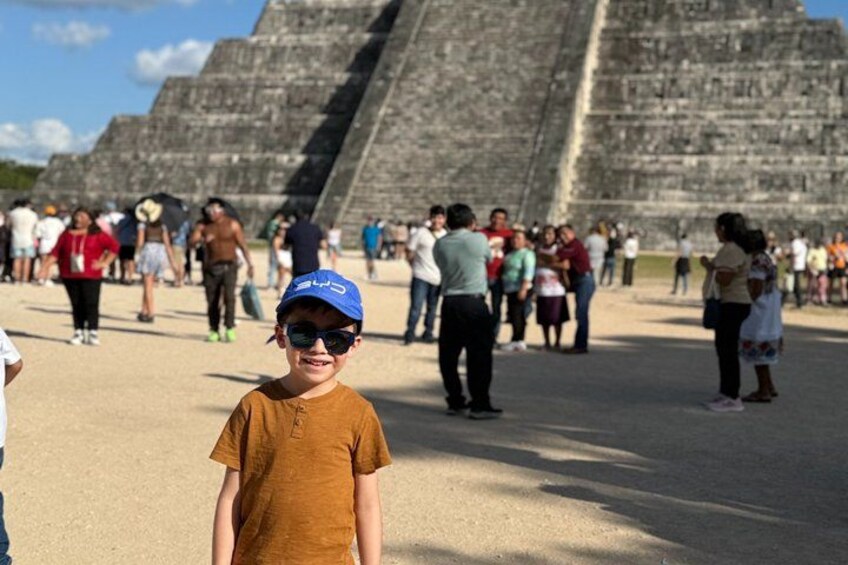 A person in a brown shirt and blue cap standing in front of ancient stone pyramids with tourists in the background. 4