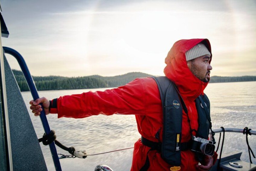 A person in a red jacket on a boat, holding onto a blue railing, with a body of water and hills in the background. 5