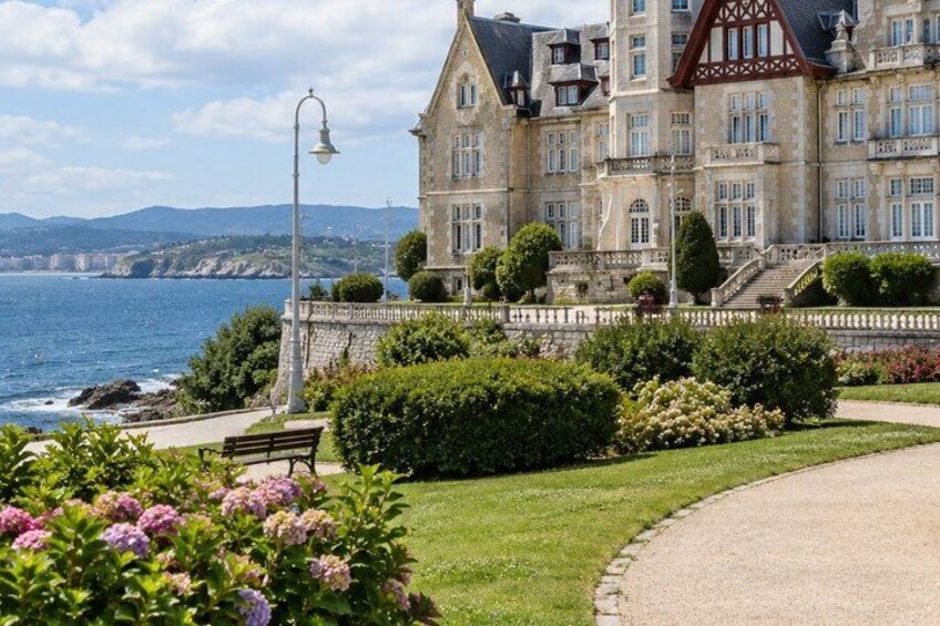 A large stone building with a balcony overlooking the sea, surrounded by well-maintained gardens and a paved walkway. 3