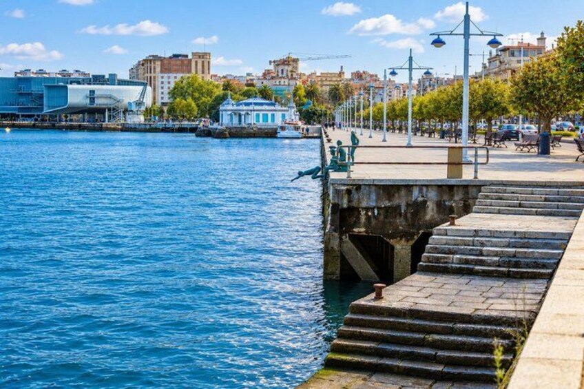 A waterfront promenade with a stone staircase leading down to the water, a blue building, and a row of trees. 2