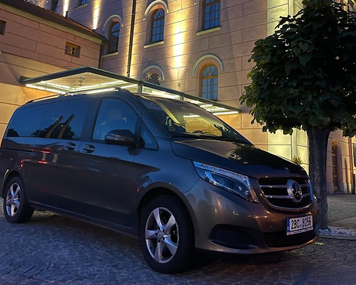 A dark-colored minivan parked in front of a building with arched windows and a tree to the right. 1