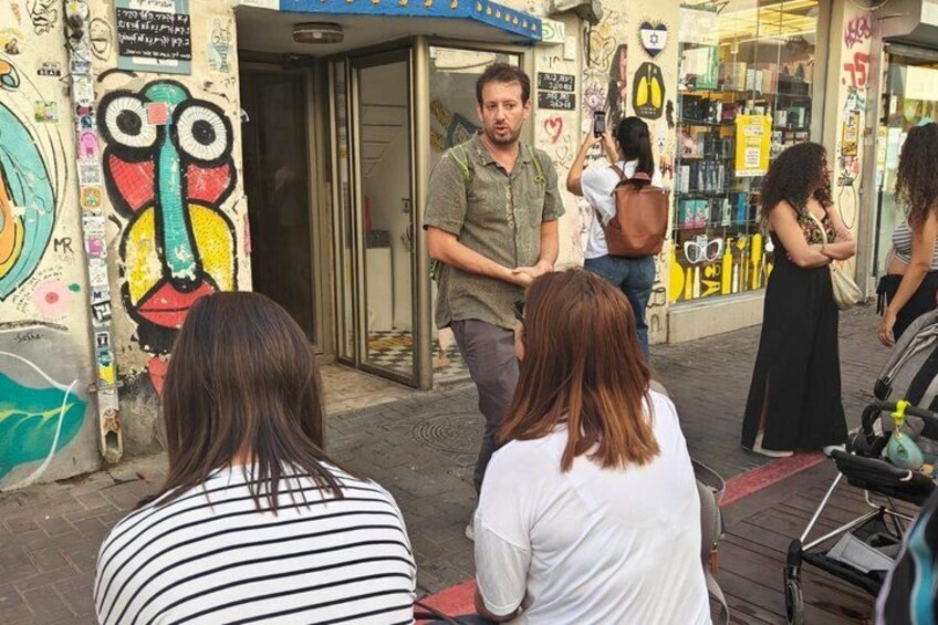 A street scene with a man walking past a shop, people standing on the sidewalk, and colorful graffiti on the walls. 2