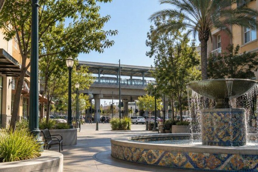 A plaza with a decorative fountain, palm trees, and a pedestrian bridge in the background. 5