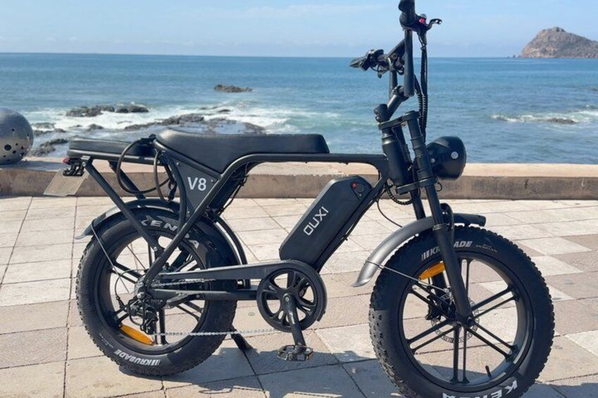 A black electric bike with the brand name 'GLUXI' parked on a tiled surface by the sea. 1