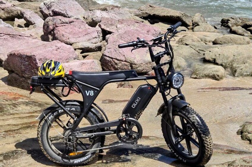 A black electric bike with a yellow helmet on the seat, parked on a rocky beach. 3