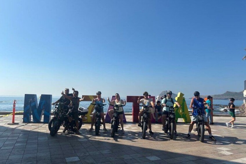 A group of cyclists and motorcyclists gathered on a paved area near a body of water, with large colorful letters spelling out a word. 5