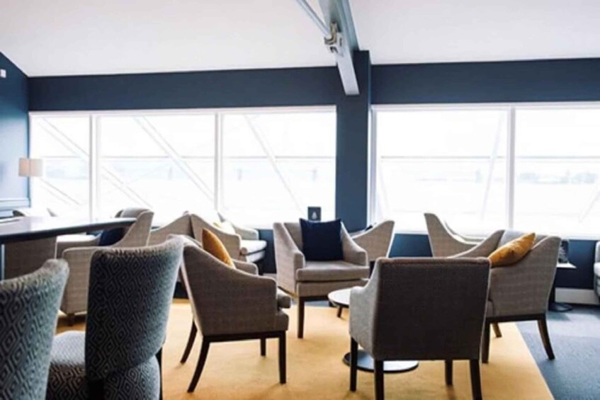 A modern lounge area with sofas and armchairs, large windows, and a carpeted floor. 3