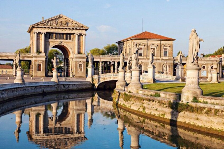 A classical building with an arched entrance, a bridge, and statues reflecting in a body of water. 1