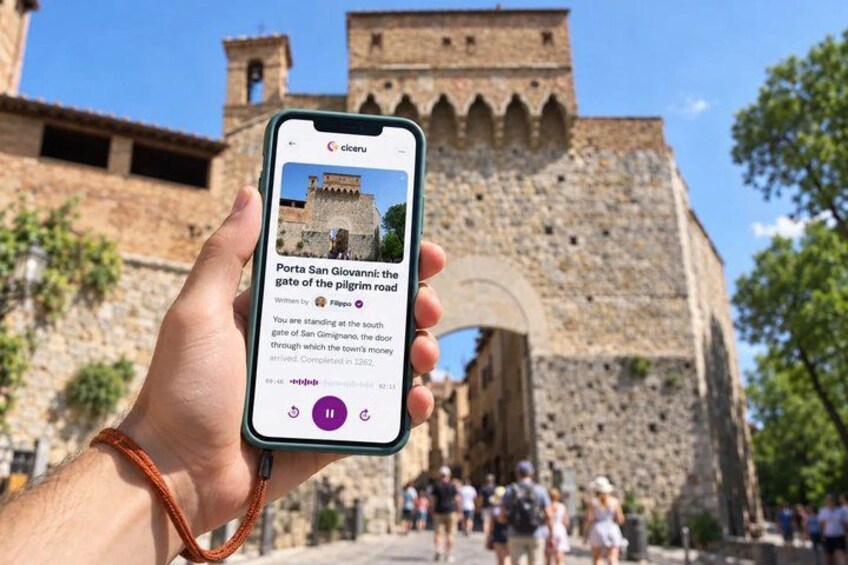 A hand holding a smartphone displaying a photo of a historic gate with a stone wall and tower in the background. 1