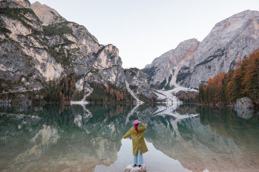 A person in a yellow jacket and pink hat standing by a clear lake with mountains in the background. 4