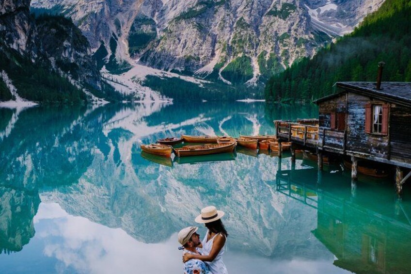 A wooden cabin by a turquoise lake with boats, surrounded by mountains. 1