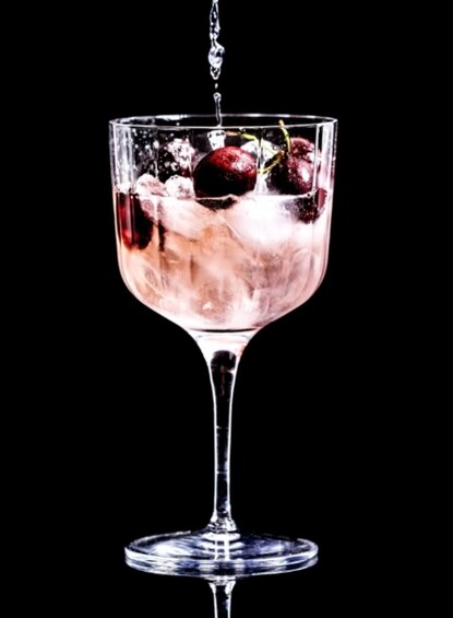 A cocktail glass with ice cubes and cherries being filled with a dark liquid. 3