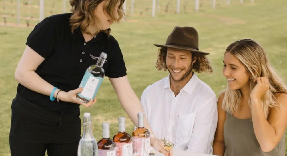 A person holding a bottle, a man in a hat, and two women at a table with drinks. 1