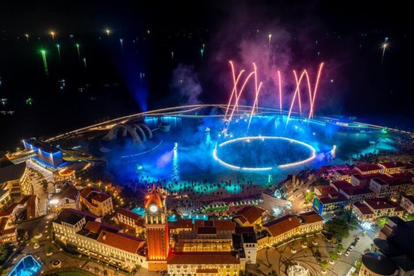 A nighttime view of a city with a large circular water feature, illuminated fountains, and fireworks. 1