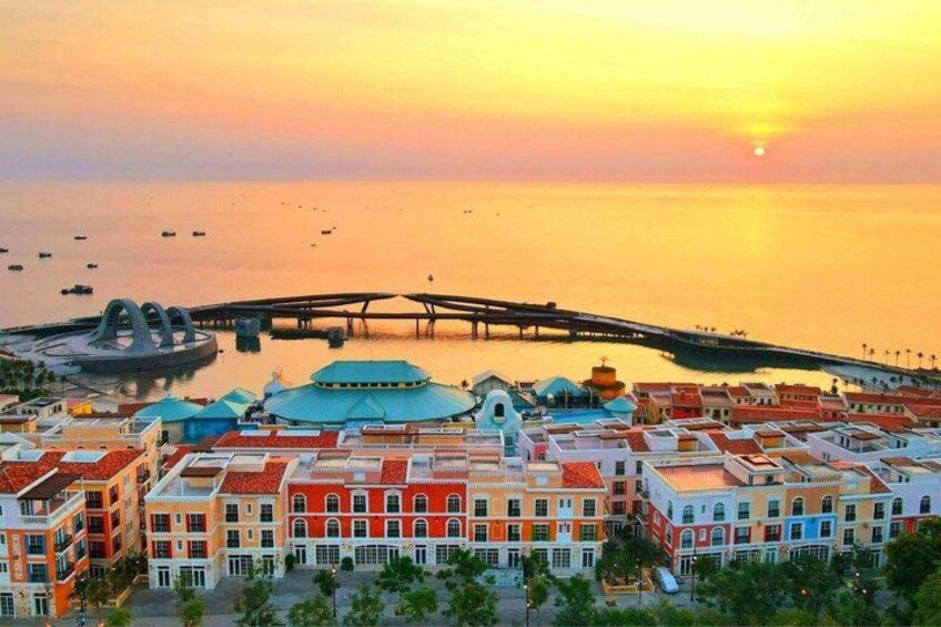 A coastal cityscape at sunset with colorful buildings, a bridge, and a circular structure. 2