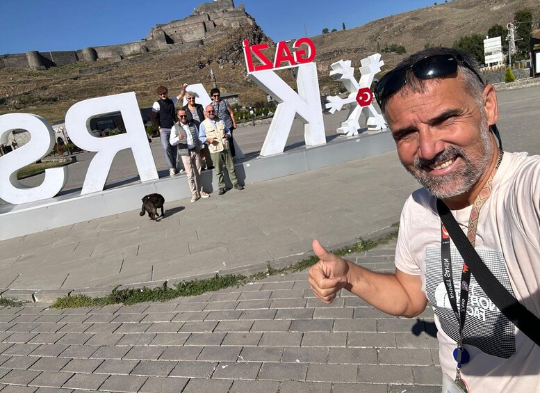 A man giving a thumbs-up gesture in front of a large white sign with the letters 'KRE' and a red 'C' on it. 4