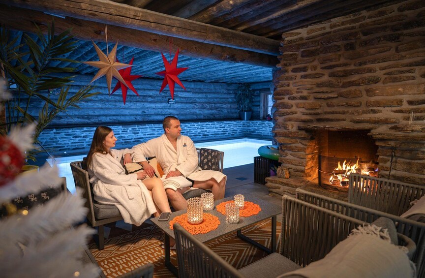 A couple in bathrobes sitting by a stone fireplace with a pool in the background. 3