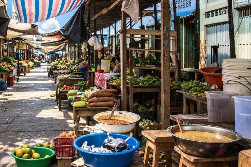 An outdoor market with wooden stalls, various fruits and vegetables, and large cooking pots. 3