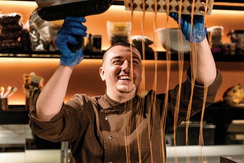 A person wearing a brown shirt and blue gloves, holding a long noodle above a kitchen counter. 1
