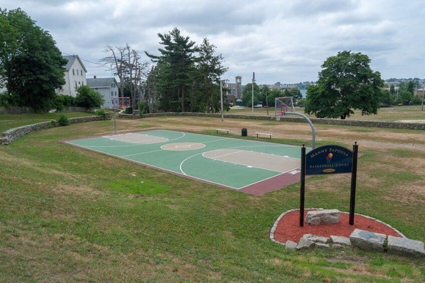 A basketball court with a sign reading 'HANNY PAPOLA BASKETBALL COURT' surrounded by trees and a house in the background. 1