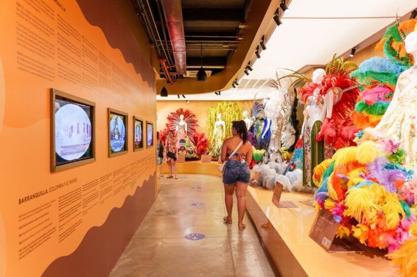 An indoor exhibition with framed photographs on the wall, a central display of colorful feathered decorations, and a person observing the exhibits. 5