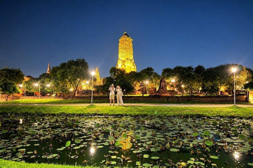 A night scene with a illuminated temple, a pond with lily pads, and two people standing near the temple. 1