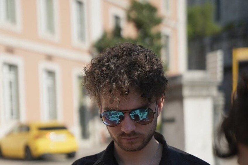 A person with curly hair and sunglasses, standing outdoors with buildings and a yellow car in the background. 3