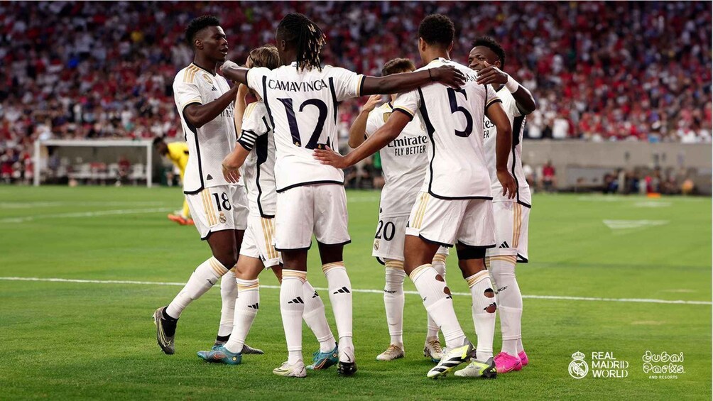 A group of soccer players in white jerseys with numbers and names, celebrating on a field with a crowd in the background. 3
