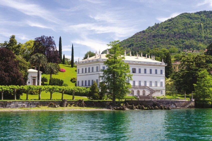 A large white building with a green lawn and trees, surrounded by water and hills. 2
