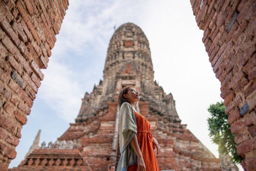 A person in traditional orange robes standing between two brick structures with a large, ancient temple in the background. 1