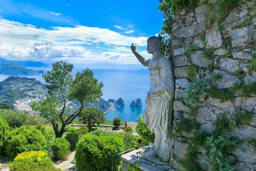 A statue of a woman overlooking a coastal landscape with a view of the sea and distant mountains. 1