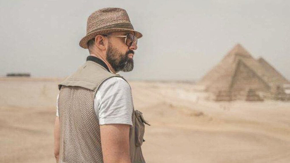 Man with hat poses with Pyramids of Giza in the distance